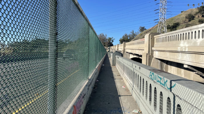 A narrow alley in Los Angeles. To its left is a screen protecting pedestrians from the highway. To its right is the LA River Bridge.