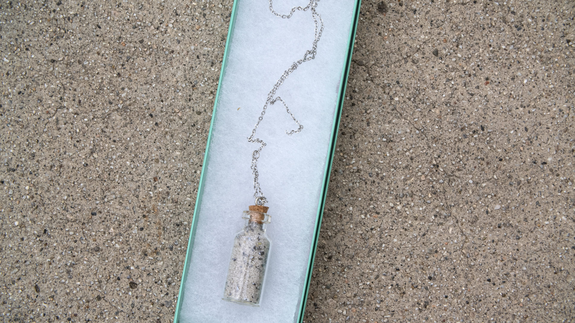 A small glass souvenir bottle filled with sand and bits of metal, on a silver chain, in a sea-foam green box, over concrete. 