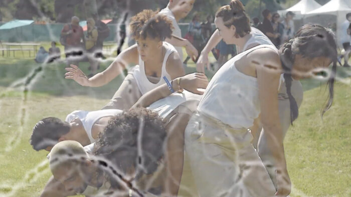 Video still of a group of people in white tank tops and beige bottom garments moving together in a cluster.