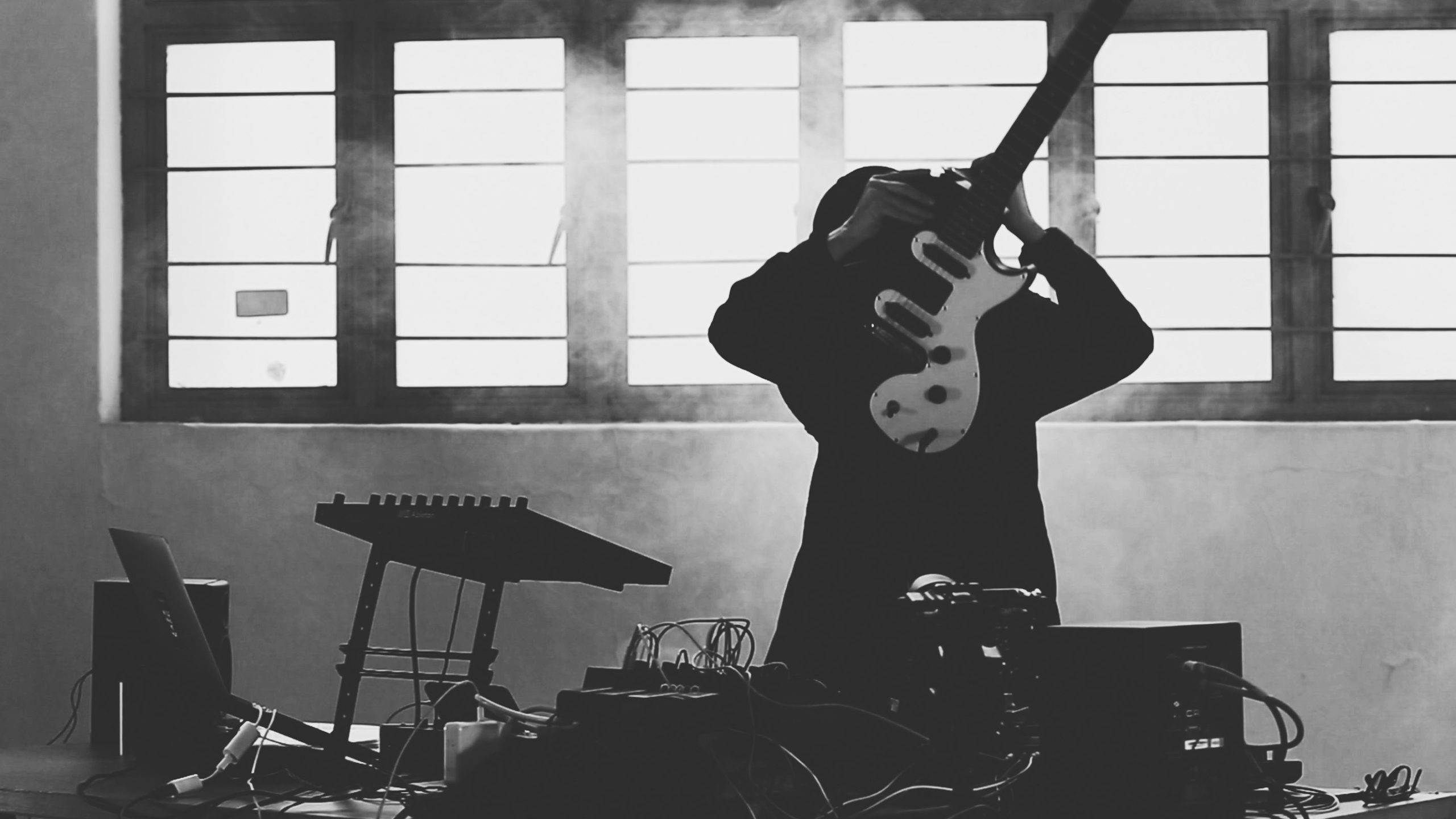 Woman holdfing an electric guitar in front of her face, standing in front of a table with various electronic equipment