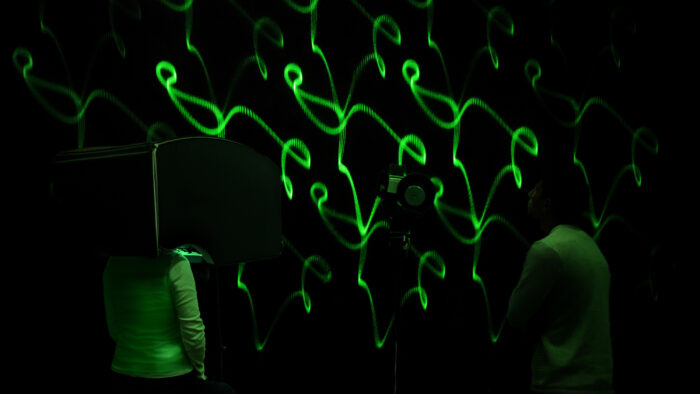 Two people stand in front of a wall on which green lasers make twisting shapes. The person on the left stands with their head inside of a console. The person on the right looks at the wall.